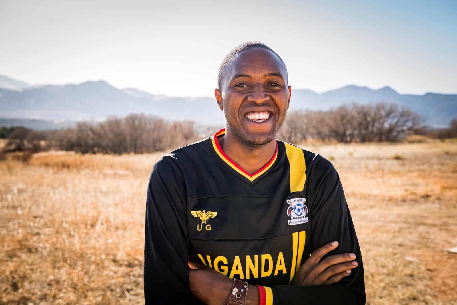 A man wearing a black jersey that says Uganda crosses his arms and smiles. He stands outside with trees and mountains in the distance.