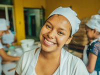 A young woman wearing a white chef's hat, white shirt and apron smiles at the camera, standing in a bakery.