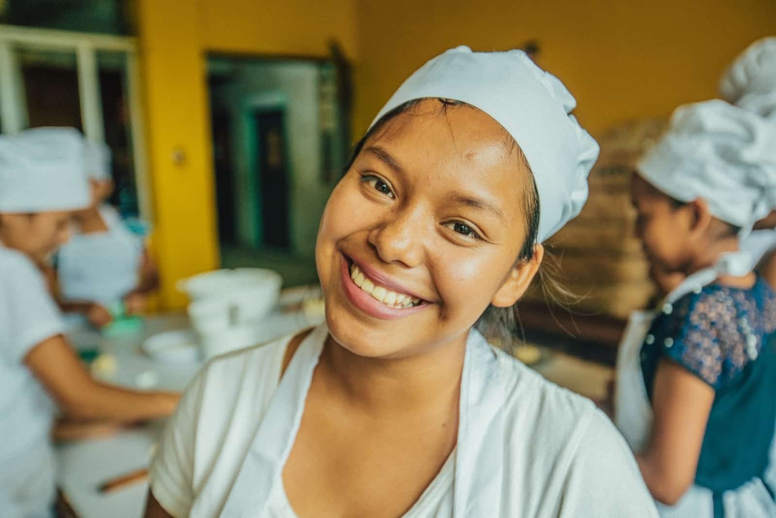 A young woman wearing a white chef's hat, white shirt and apron smiles at the camera, standing in a bakery.