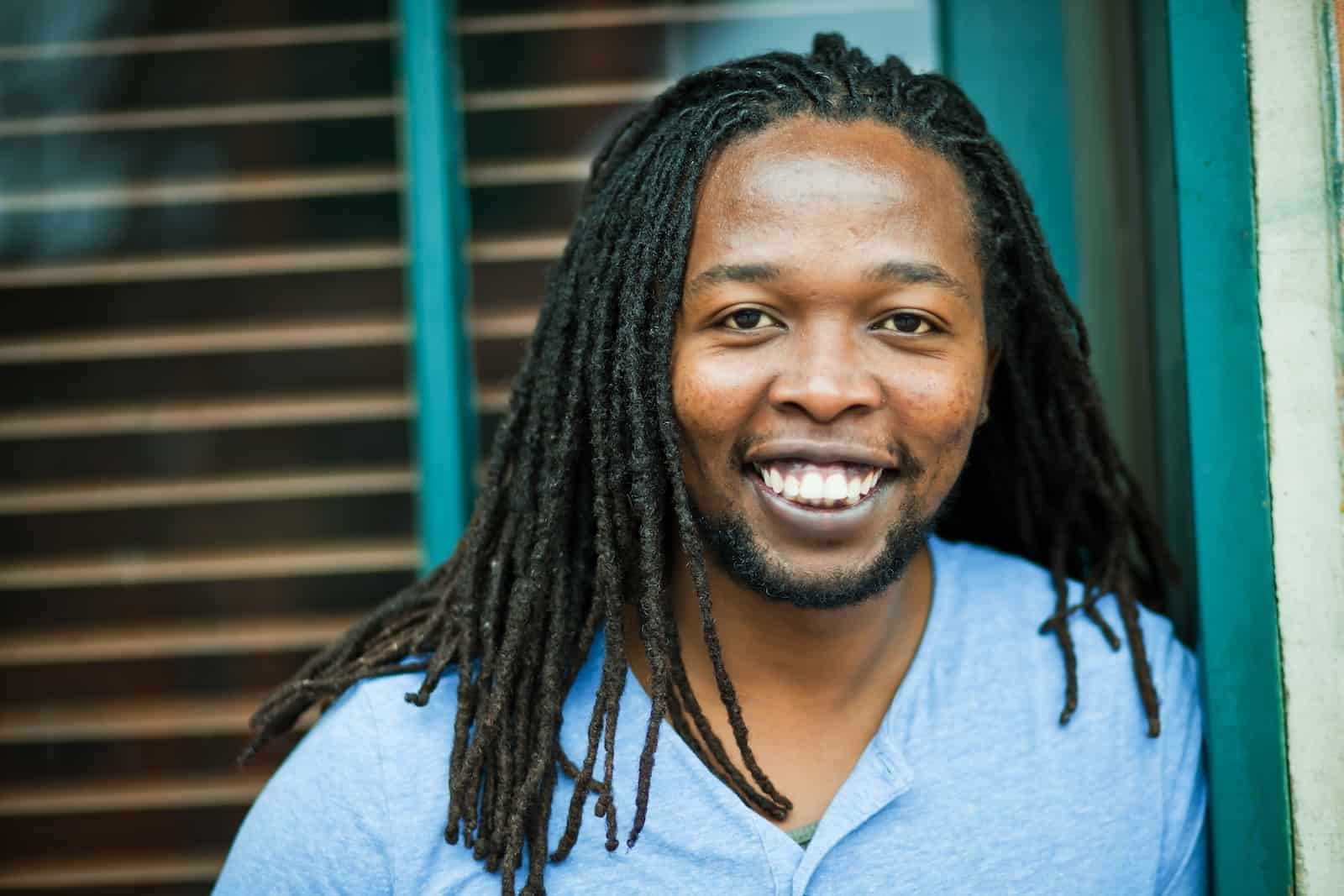 A man in a blue shirt stands outside in front of a window and smiles at the camera.