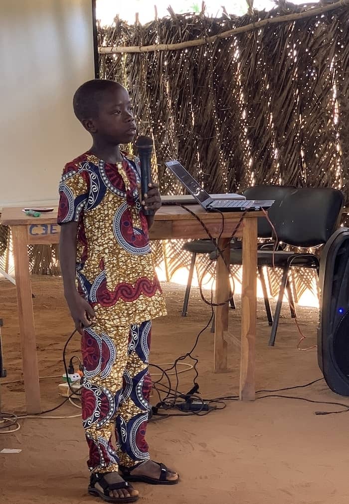 A boy in a patterned yellow and red Togolese outfit stands, speaking into a microphone. He is in a room made from woven leaves. He is an example of the Easter message of hope.