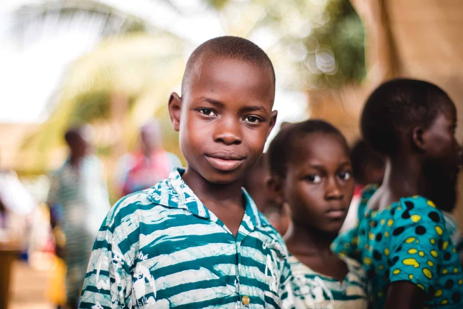 A boy from Togo wearing a green and white shirt looks into the camera. He is surrounded by children wearing similar colors. They stand outside with palm trees in the background.