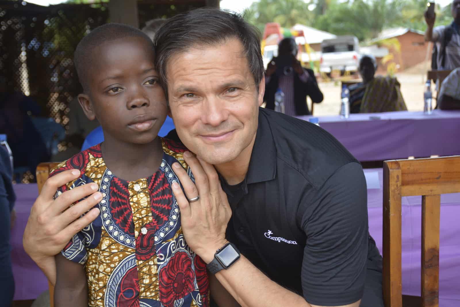 A man, Santiago "Jimmy" Mellado, wearing a black shirt that says "Compassion" stoops down and puts his arms around a young boy wearing a yellow and red Togolese shirt. 