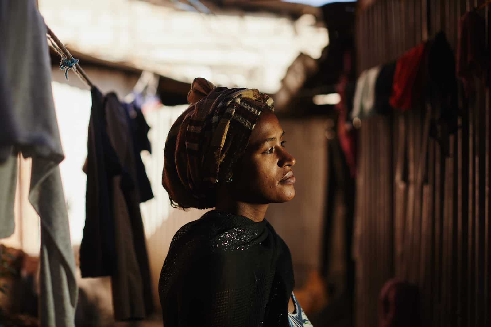 A woman wearing a plaid headscarf and black shawl looks to the side. She stands in a dark alley with laundry hanging on the line.