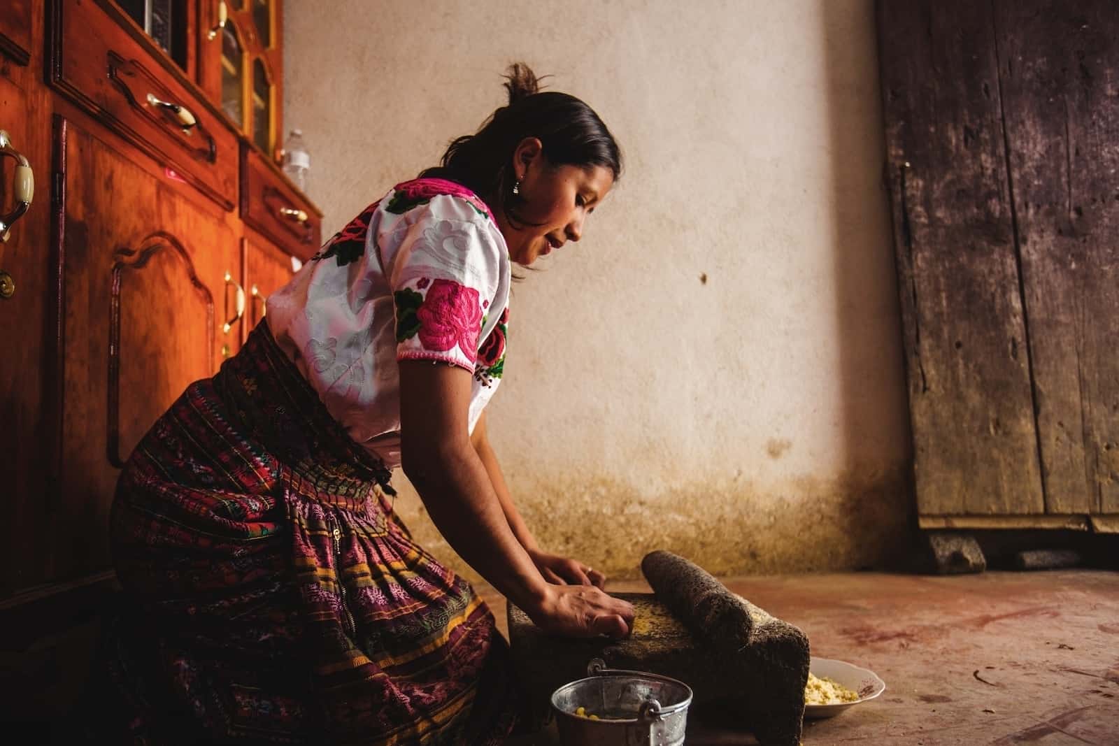 A woman wearing a colorful skirt with a pink and white shirt kneels in a room, grinding corn on a stone.