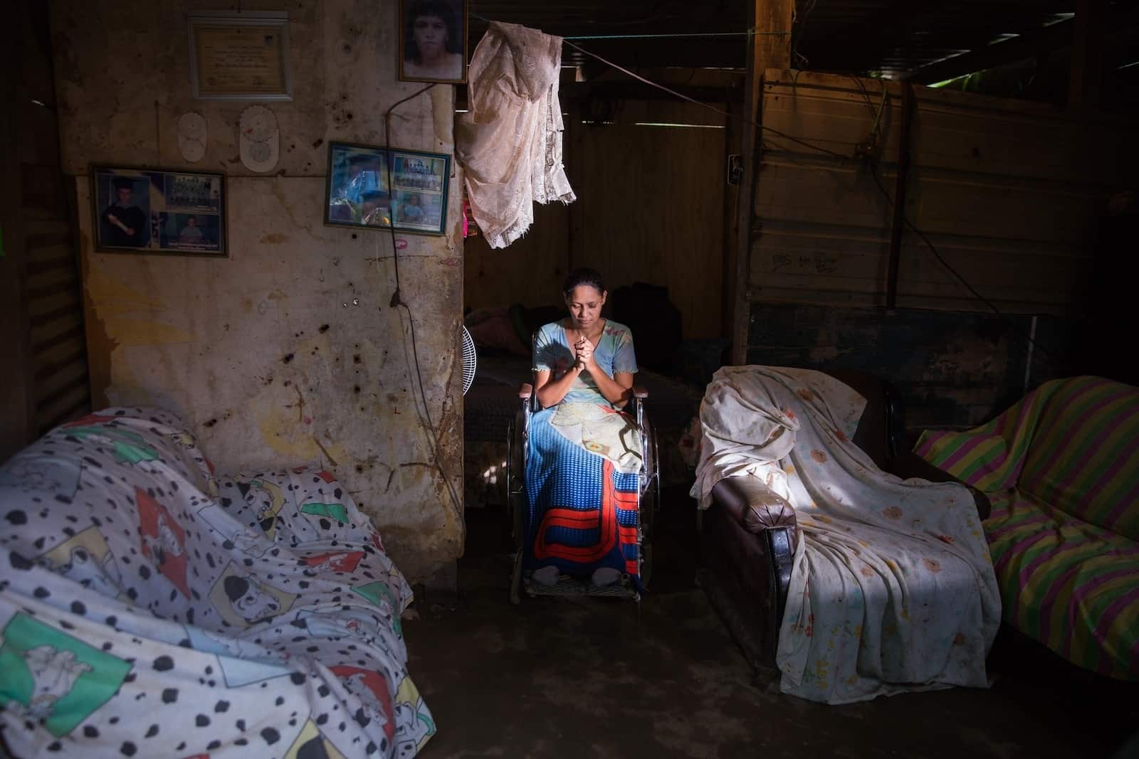 A woman sits in a wheelchair with her head bowed and hands in the praying position. She is in a dark room with two beds on either side of her.