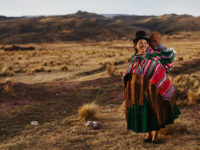 A woman wearing a black bowler hat, colorful shawl and traditional Bolivian clothes stands in a wide field, carrying a baby on her back.