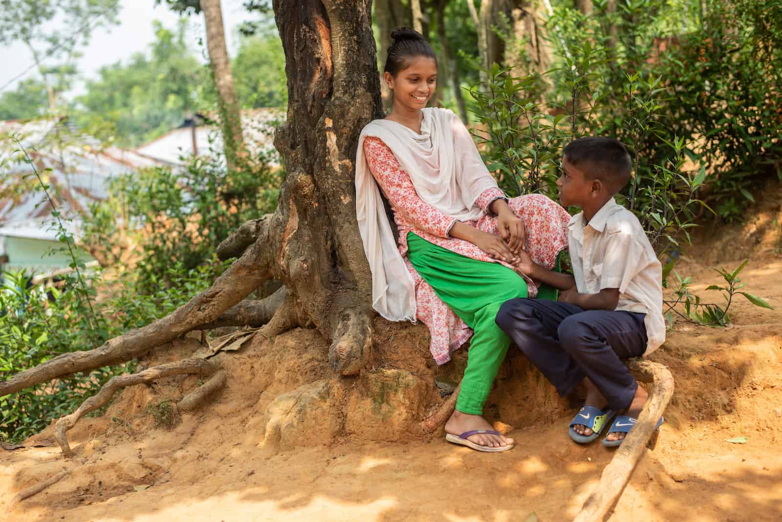 A girl wearing green pants, a pink shirt and a white scarf leans against the trunk of a large tree. A young boy wearing jeans and a white button-up shirt kneels below her as they smile at each other. 