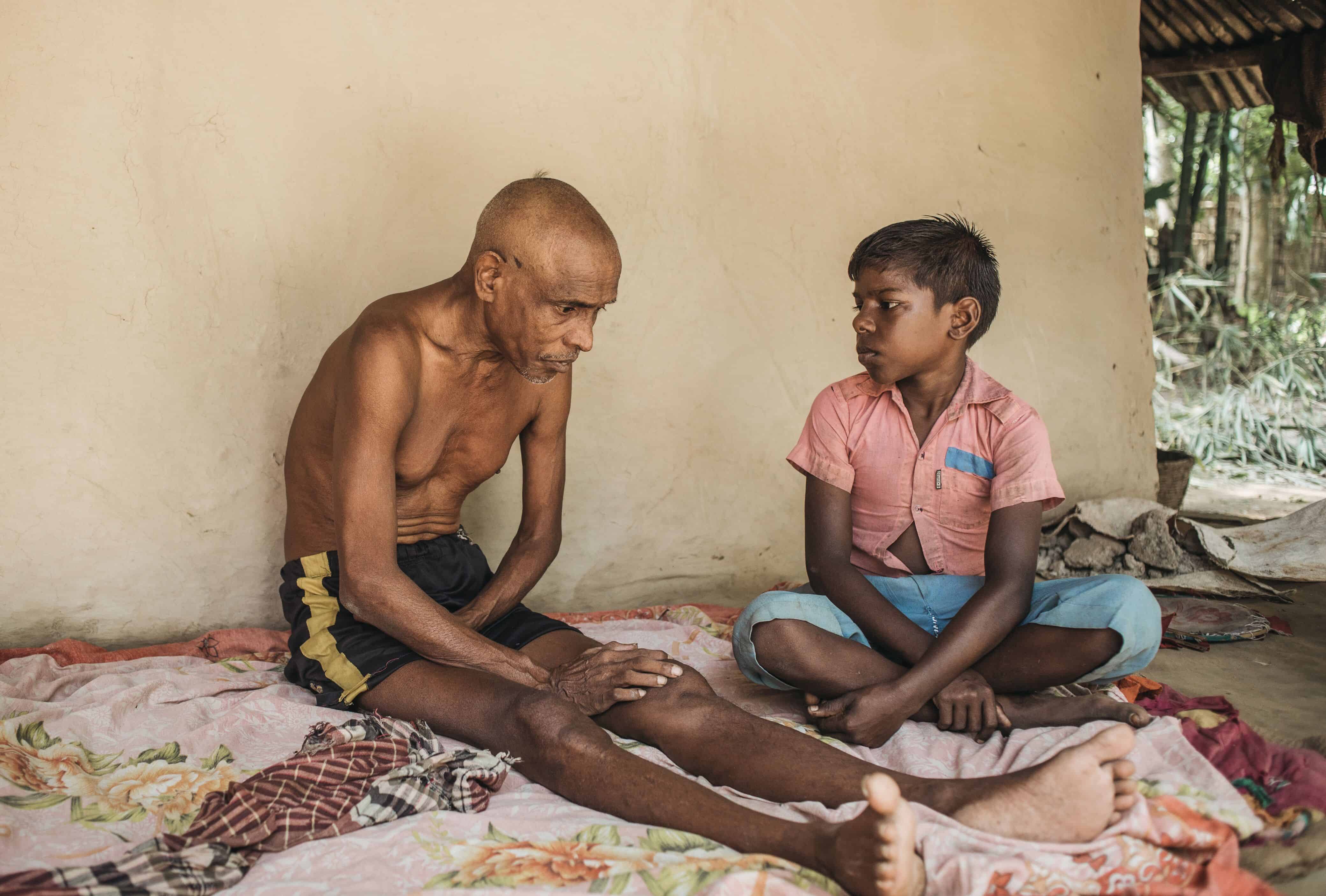 An older man without a shirt sits on a blanket against a tan colored wall, looking down at the ground. A young boy in a pink shirt sitting next to him looks at him. 