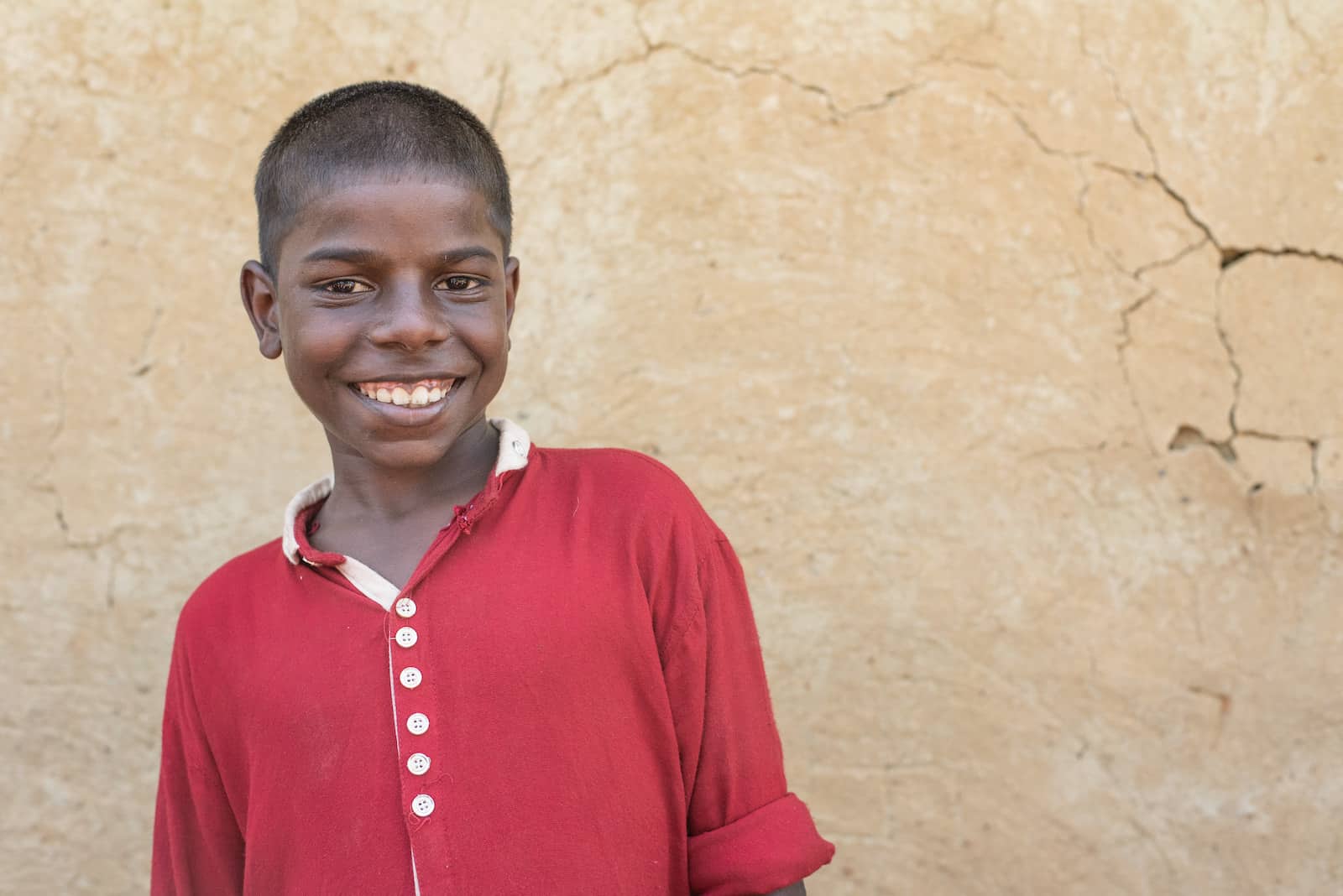 A young boy wearing a red button-up shirt smiles widely, standing in front of a tan wall. 