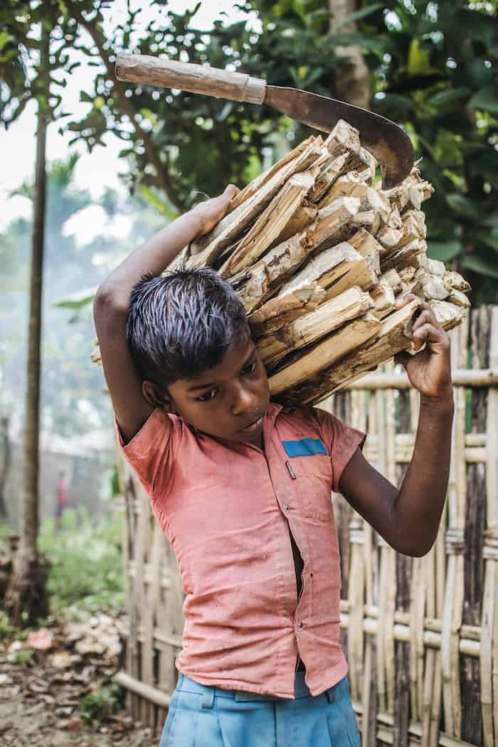 A 10-year-old boy wearing a coral-colored shirt carries a large bundle of wood on his shoulder, with a machete stuck in the top of the wood. 