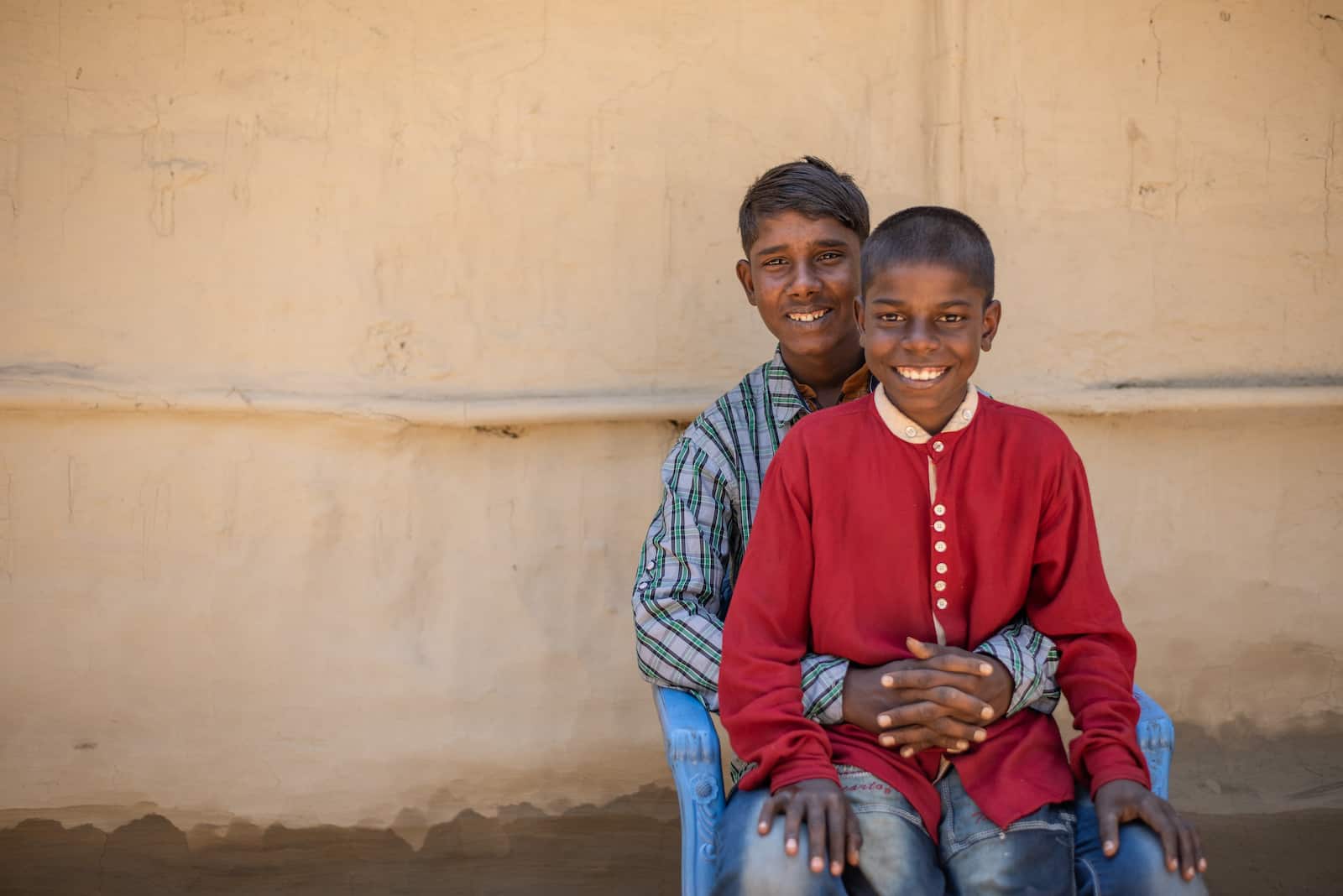 A young smiling boy wearing a red shirt sits in the lap of a smiling teenager wearing a blue plaid shirt, with his hands clasped around his brother. 