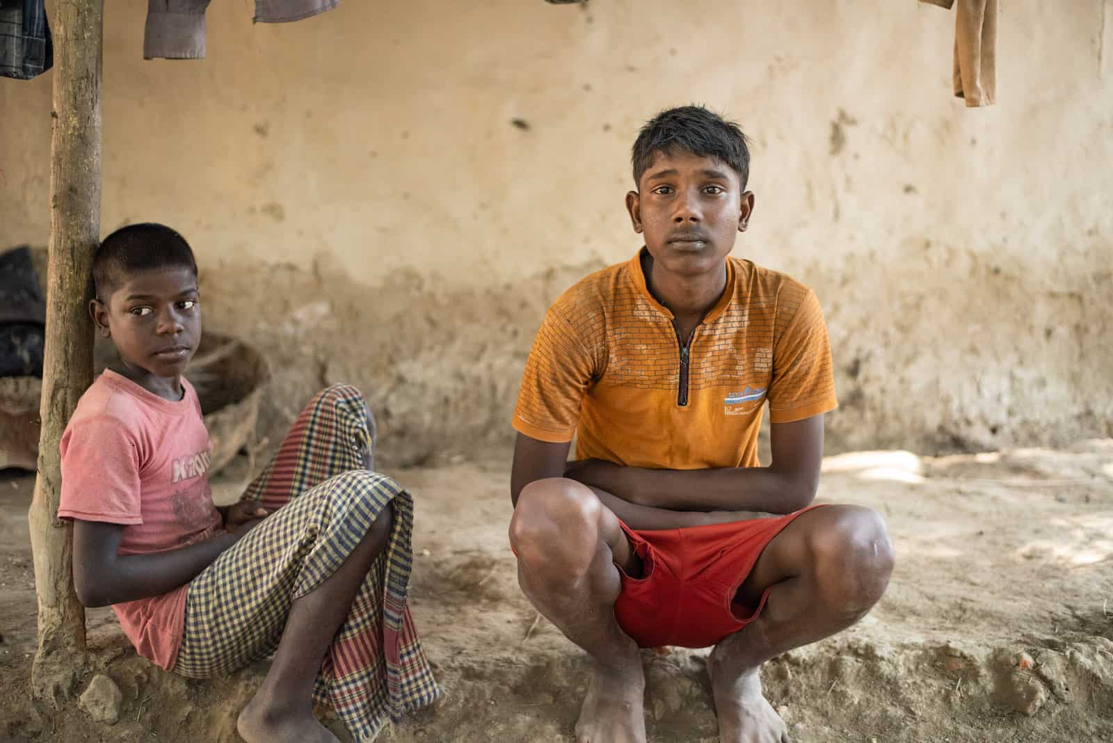 A 15-year-old boy wearing an orange shirt and red shorts crouches in front of an adobe wall. To his right is a younger boy, wearing a pink shirt and a traditional plaid skirt.