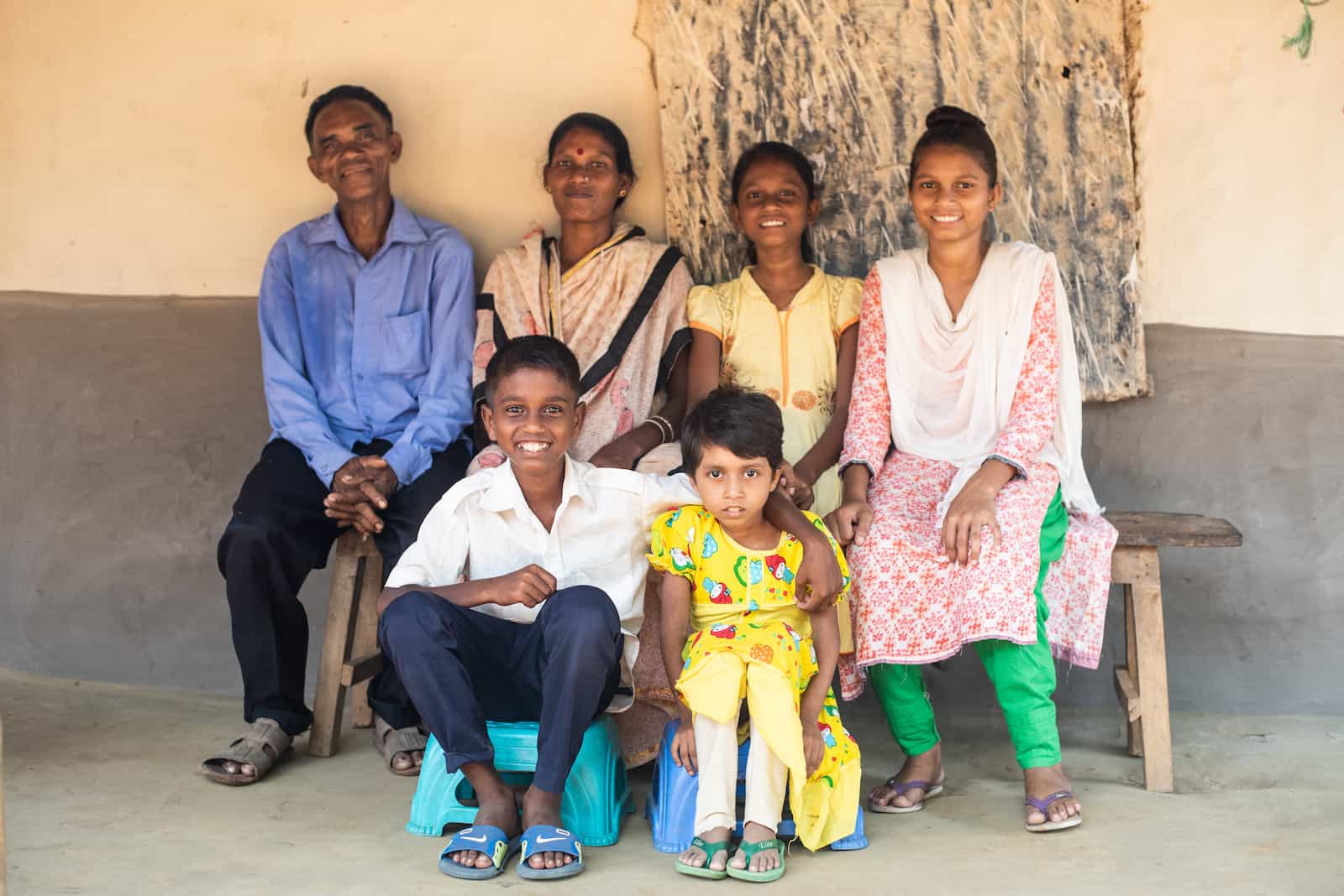 A family of six sits outside, smiling, including an older man in blue, a woman wearing a tan sari, a girl in a yellow dress, and another girl in a pink and green outfit. Below them are a young boy in a white shirt and blue pants and a young girl in a yellow dress. 