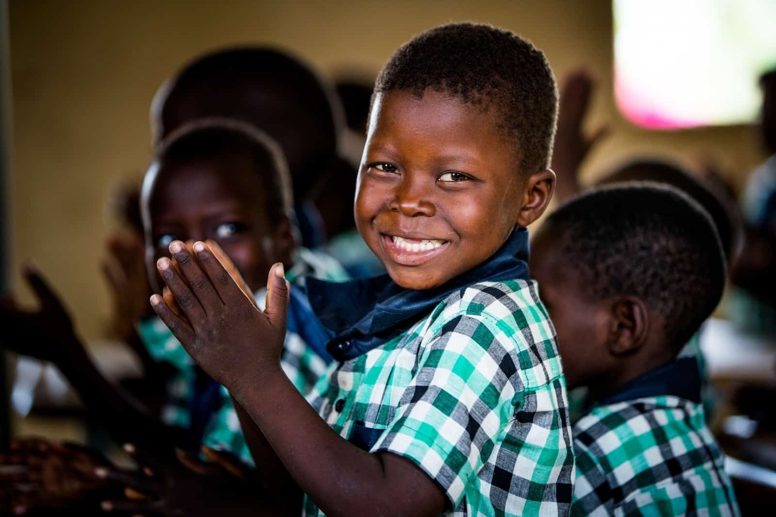 A boy in Burkina Faso wearing a green and blue checked shirt holds his hands together in praying while smiling at the camera. He is in a classroom surrounded by other children praying. 