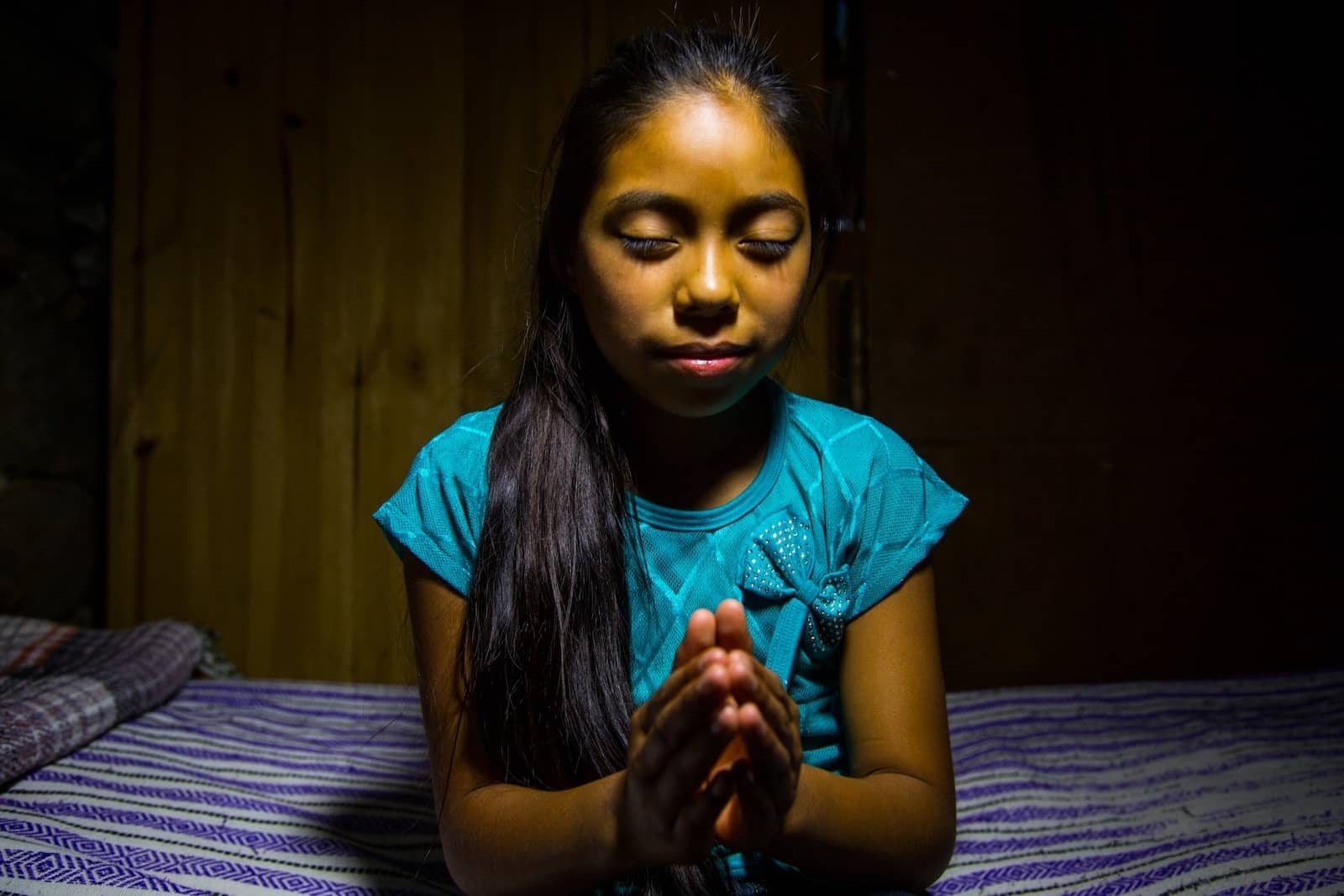 A girl in Mexico wearing a green shirt with long hair in a ponytail sits in a dark room with eyes closed and hands in the praying position. 