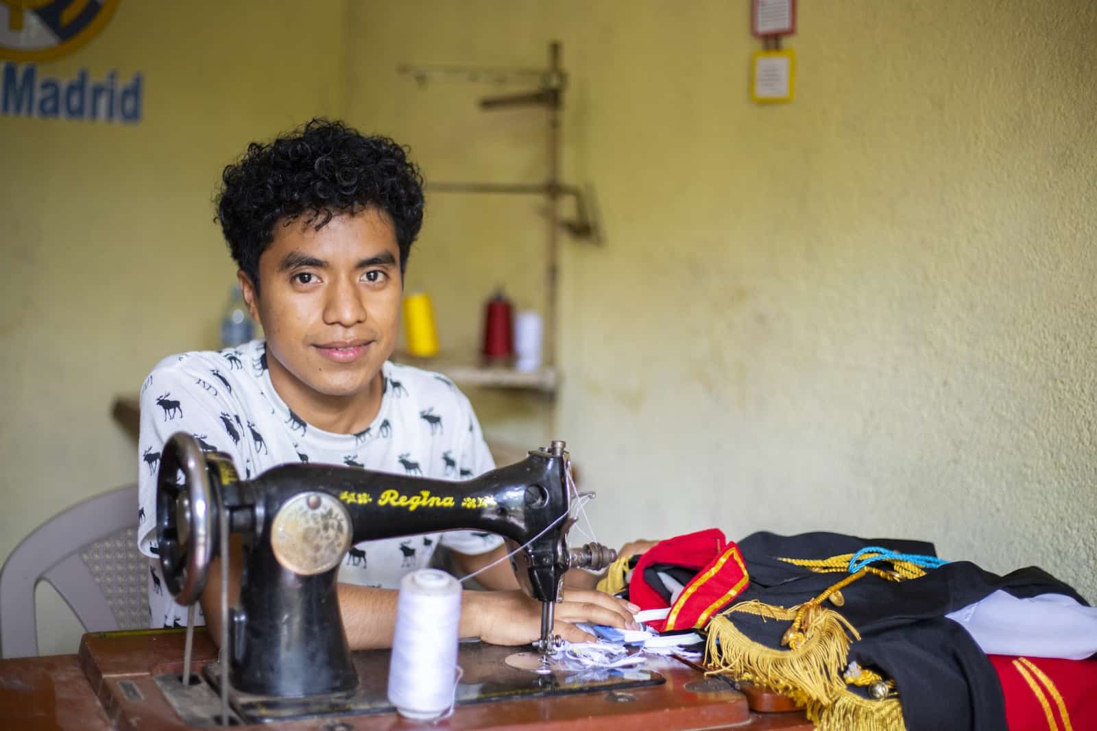 A young man sits at a table with a sewing machine in front of him and uniforms on the table. 