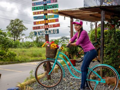 A woman in a pink shirt, jeans and a baseball cap sits on a turquoise bike on a roadside, with a sign behind her that reads, 