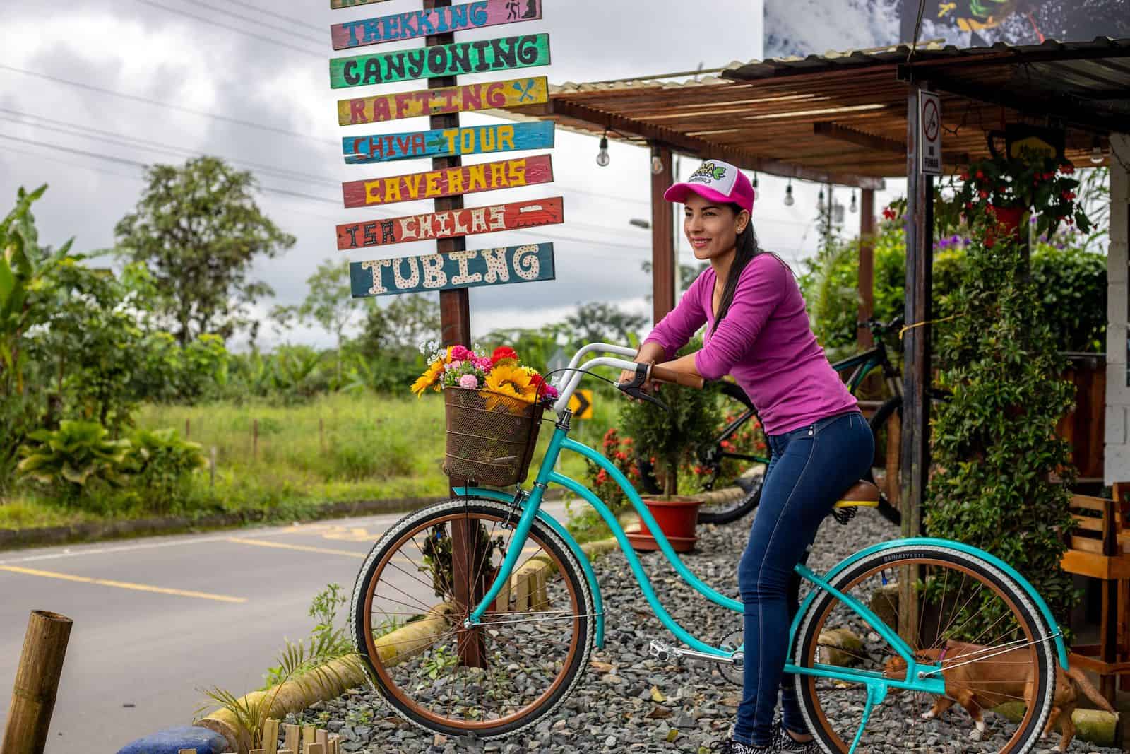 A woman in a pink shirt, jeans and a baseball cap sits on a turquoise bike on a roadside, with a sign behind her that reads, "Trekking, Canyoning, Rafting, Tubing," etc.