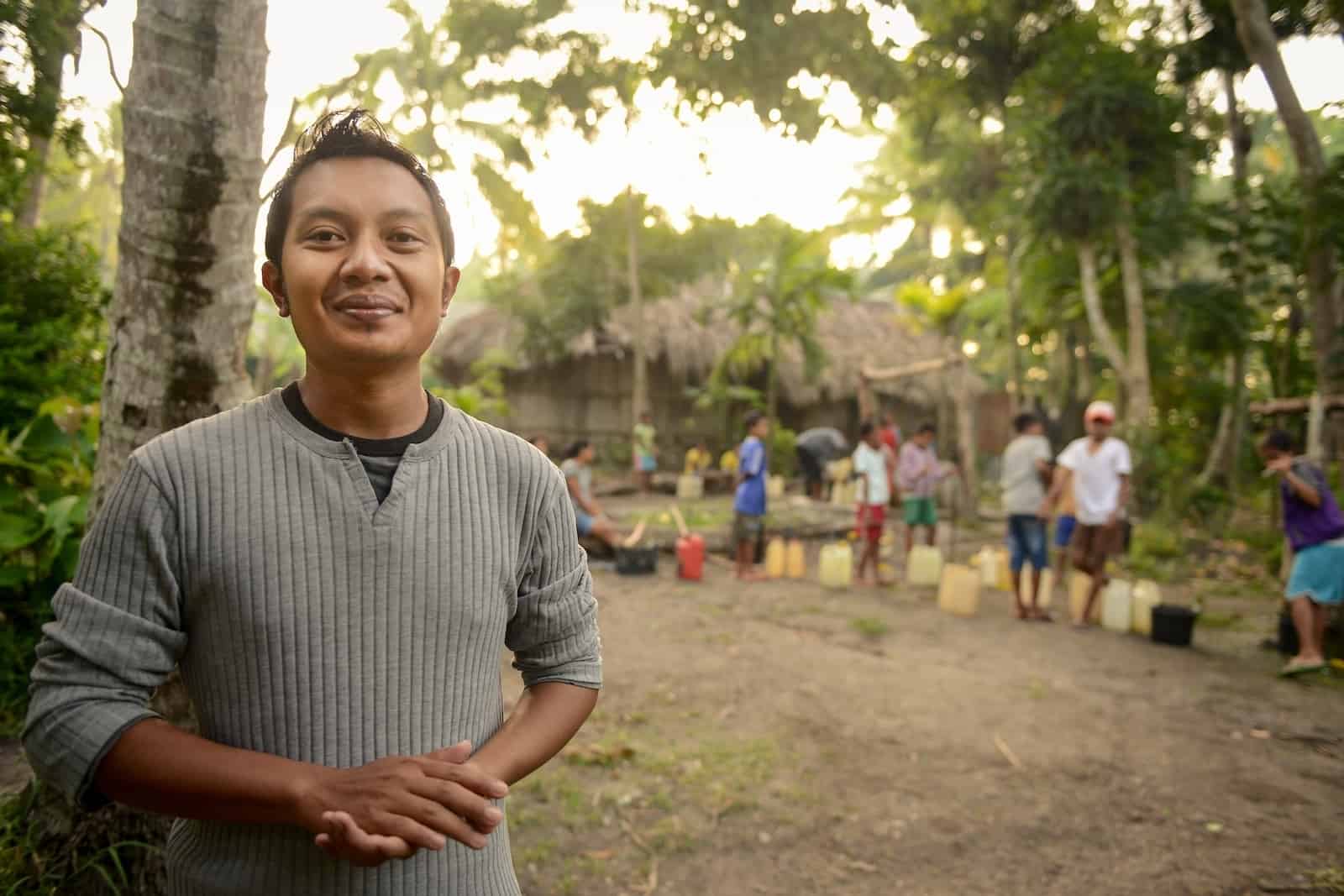 A young man who is a Compassion graduate stands outside in a gray sweater, with his hands clasped in front of him. Behind him are palm trees and a thatched building with a line of people in front of it, with jerry cans.