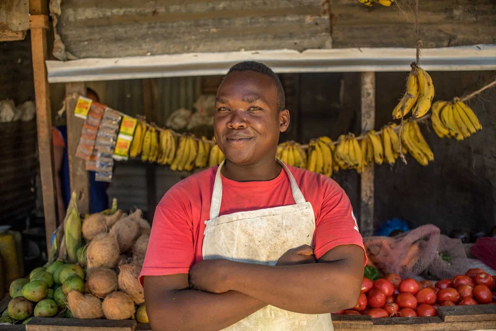 A Compassion graduate wearing a red T-shirt and white apron stands with his arms crossed, in front of a grocery stall, full of bananas, apples and coconuts. 