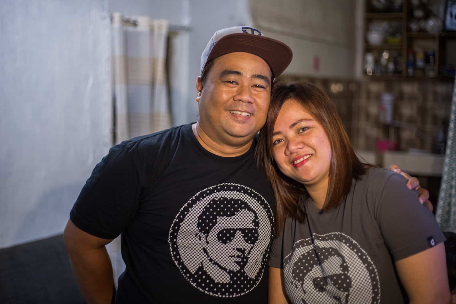 A man and a woman wearing matching black and grey T-shirts smile at the camera, the man's arm around the woman.