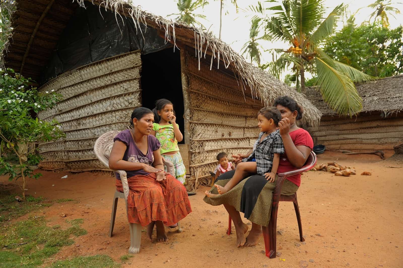 Two women sit in chairs in front of a home woven from leaves and bamboo, showing poverty in Asia. Each woman has a child next to her or on her lap.