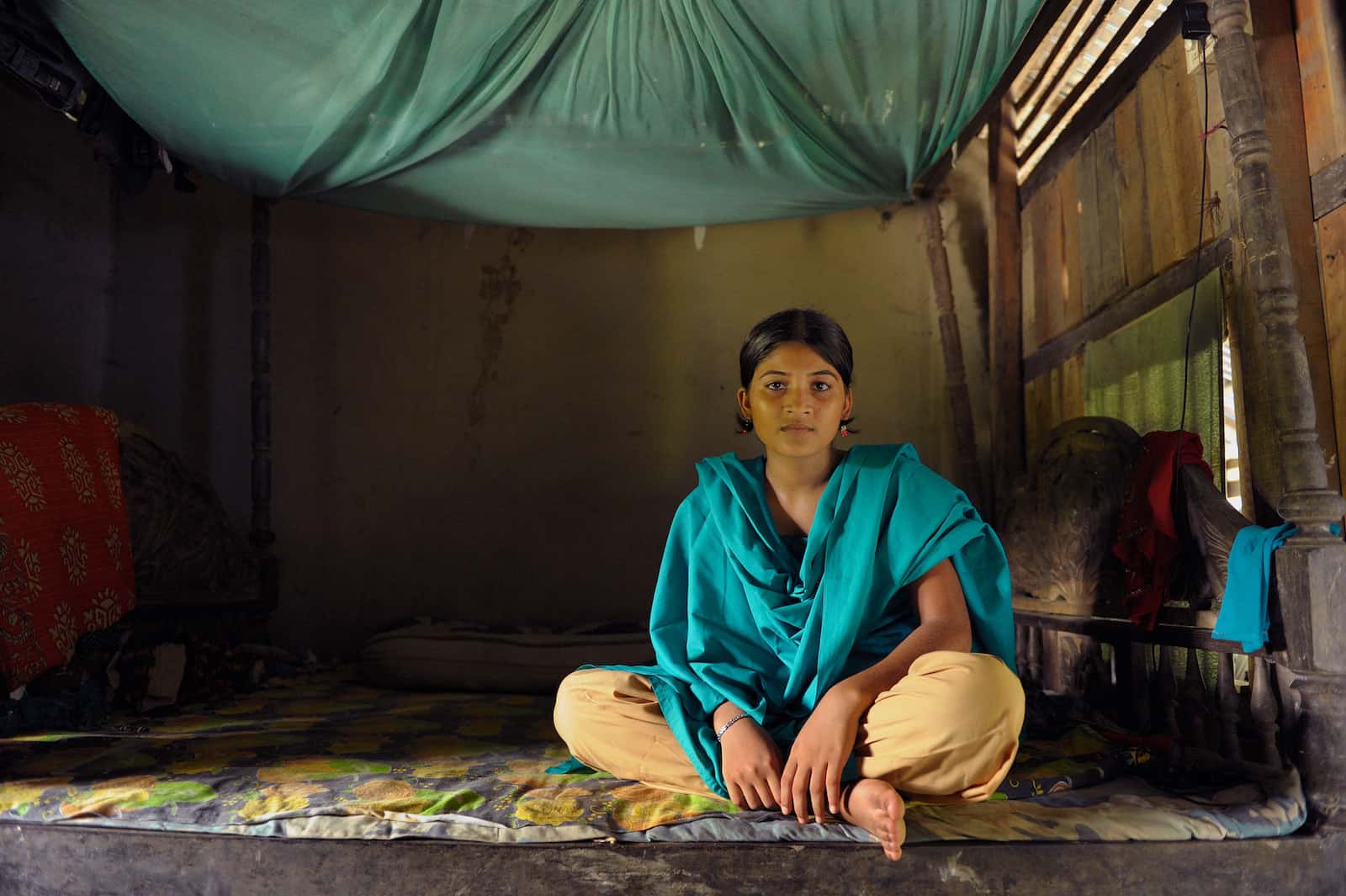 A girl in yellow pants and a green scarf sits on a woodframe bed with a mat on top and with a mosquito net above.