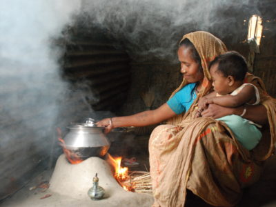 A woman with a baby in her lap sits inside in front of a small fire with a pot on top of it, with smoke in the air.