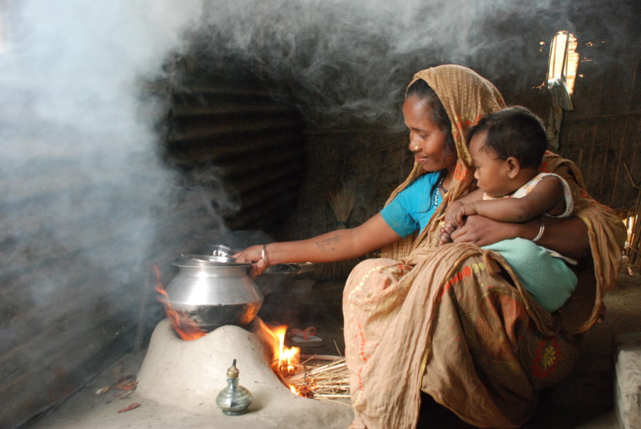 A woman with a baby in her lap sits inside in front of a small fire with a pot on top of it, with smoke in the air.