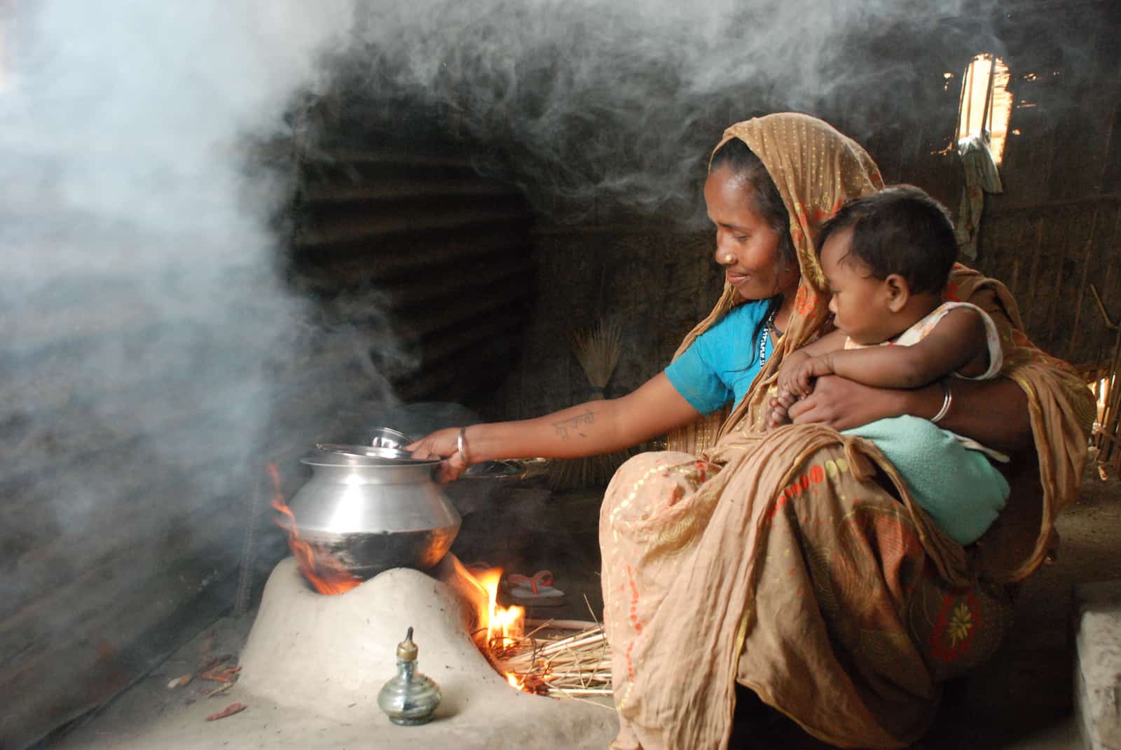 A woman with a baby in her lap sits inside in front of a small fire with a pot on top of it, with smoke in the air. 