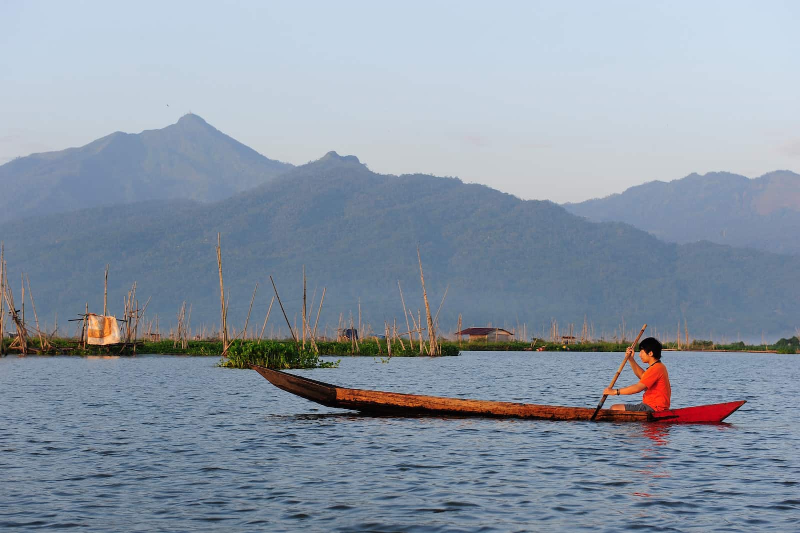 A young man in a red shirt sits in a long canoe, rowing on a large body of water, with mountains in the background.