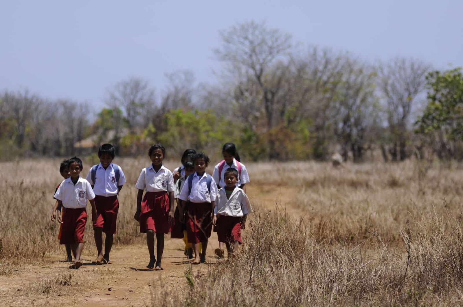 A group of students wearing red and white school uniforms walk down a dirt road in a brown field with trees in the background.
