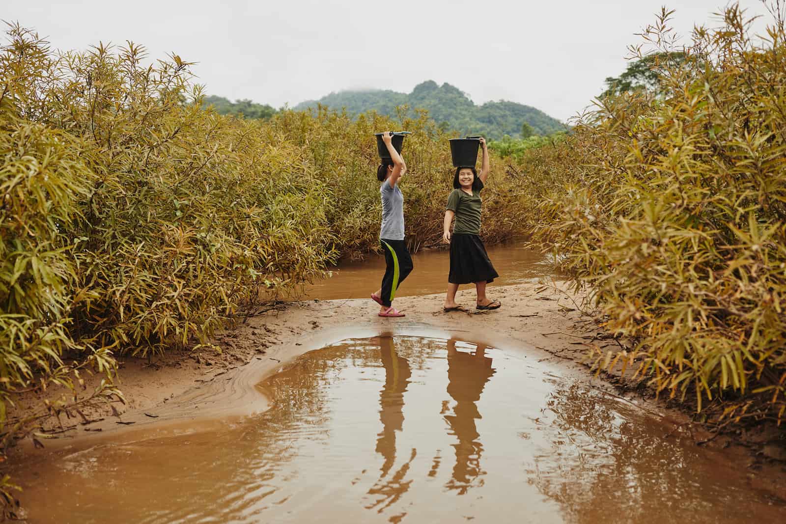 Two girls carry black buckets on their heads, standing in front of a small river, with foliage in the background.