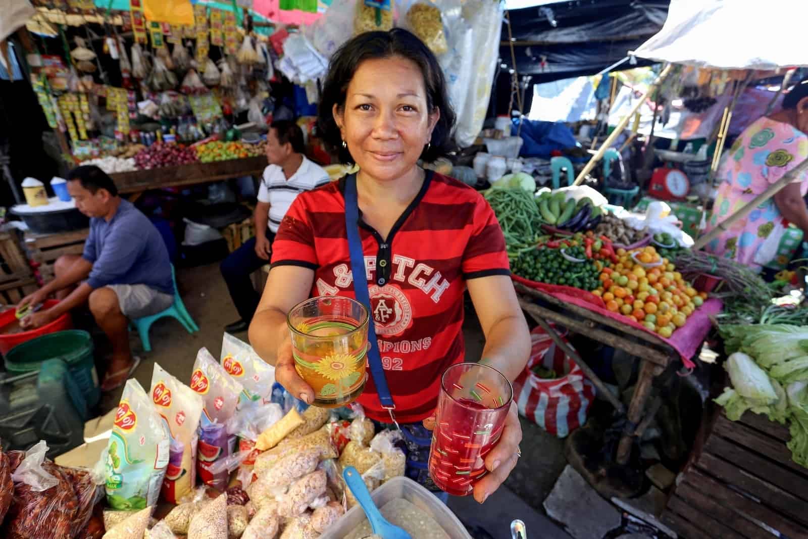A woman wearing a red shirt holds out two drinks, standing in a grocery stall.
