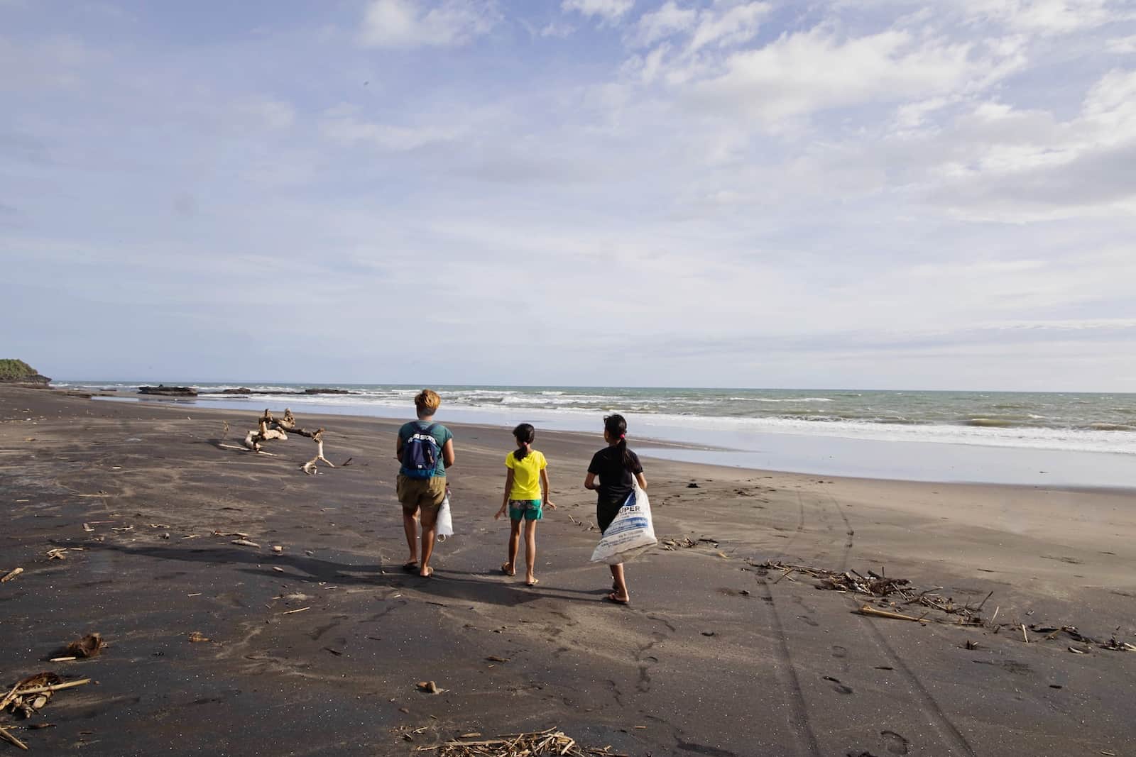 A woman walks down a wide beach with two children, holding bags in which they gather trash.
