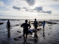 Two adults and three children stand on a beach on Bali at sunset, picking up trash.