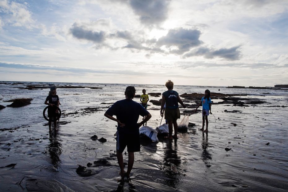 Two adults and three children stand on a beach on Bali at sunset, picking up trash.