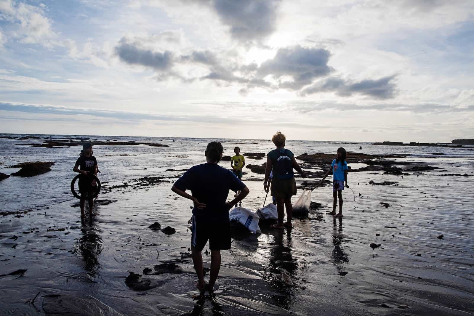 Two adults and three children who live in poverty stand on a beach on Bali at sunset, picking up trash to protect the environment. 