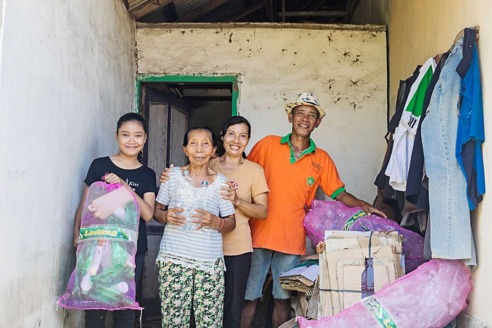 A family of a girl, grandmother, mother and father stand outside a home holding large bags full of plastic bottles. 
