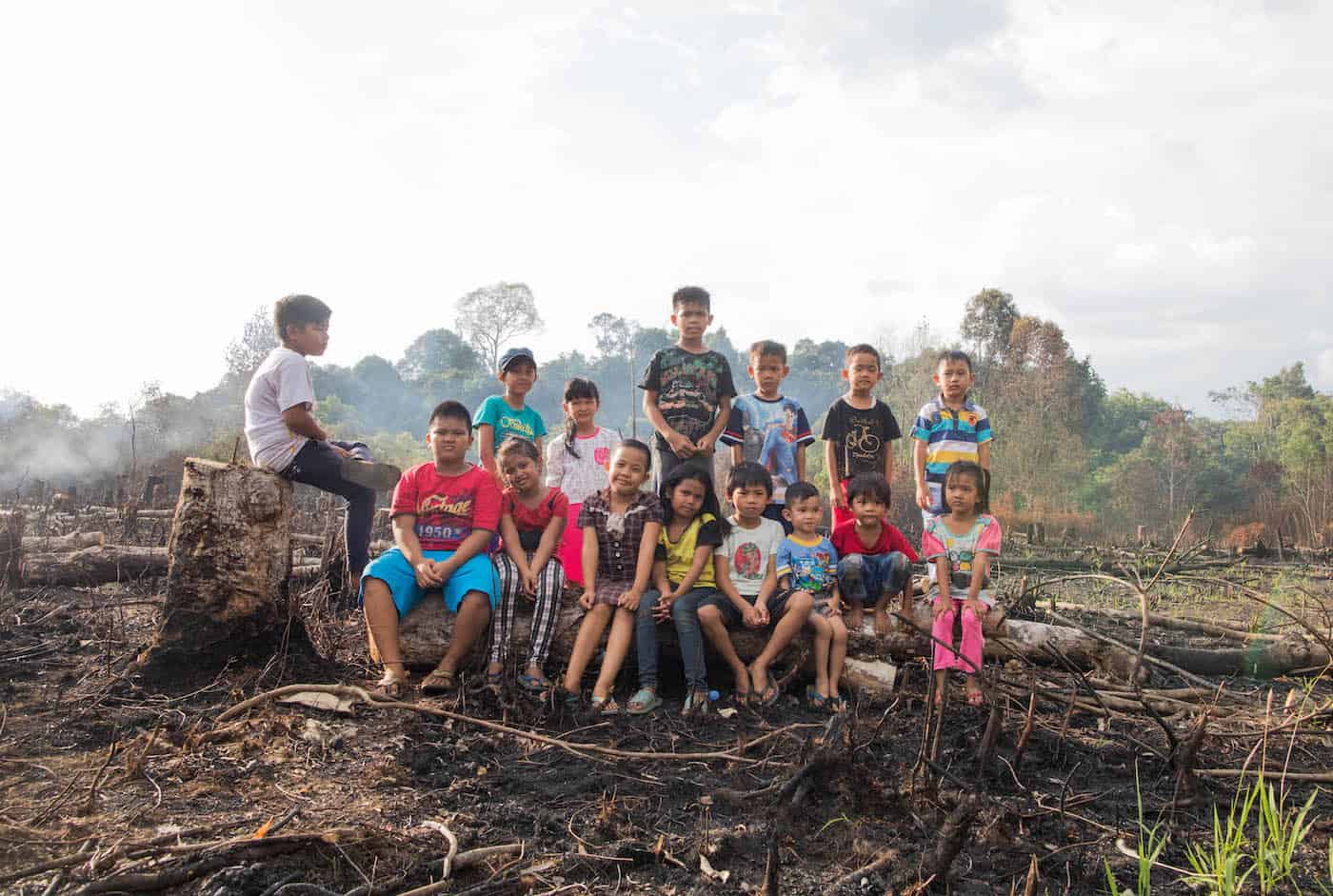 A group of children sit outside in a burned field. In the background is forest and smoke rising into the sky. 