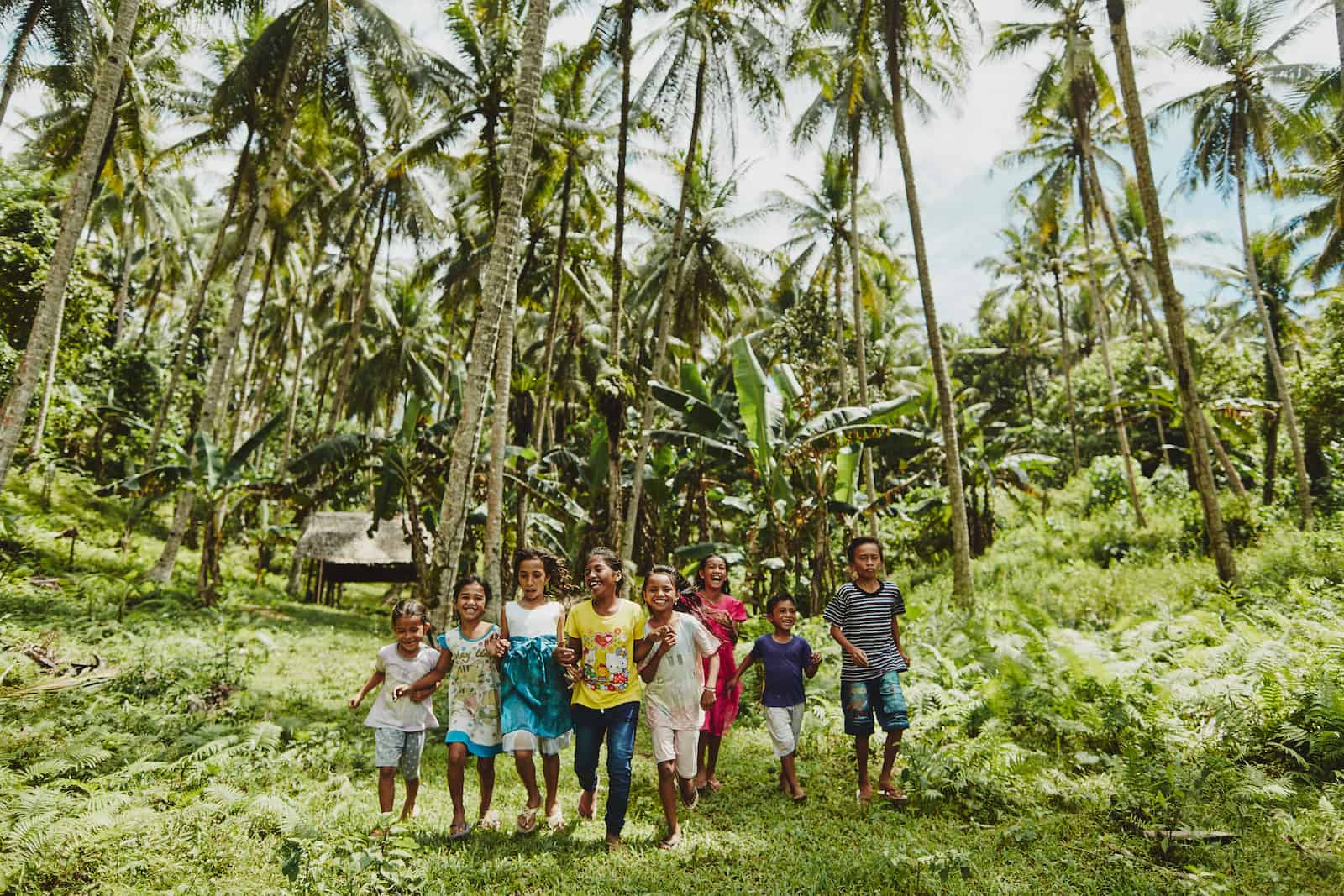A group of children run through green grass in a forest of palm trees in Indonesia.