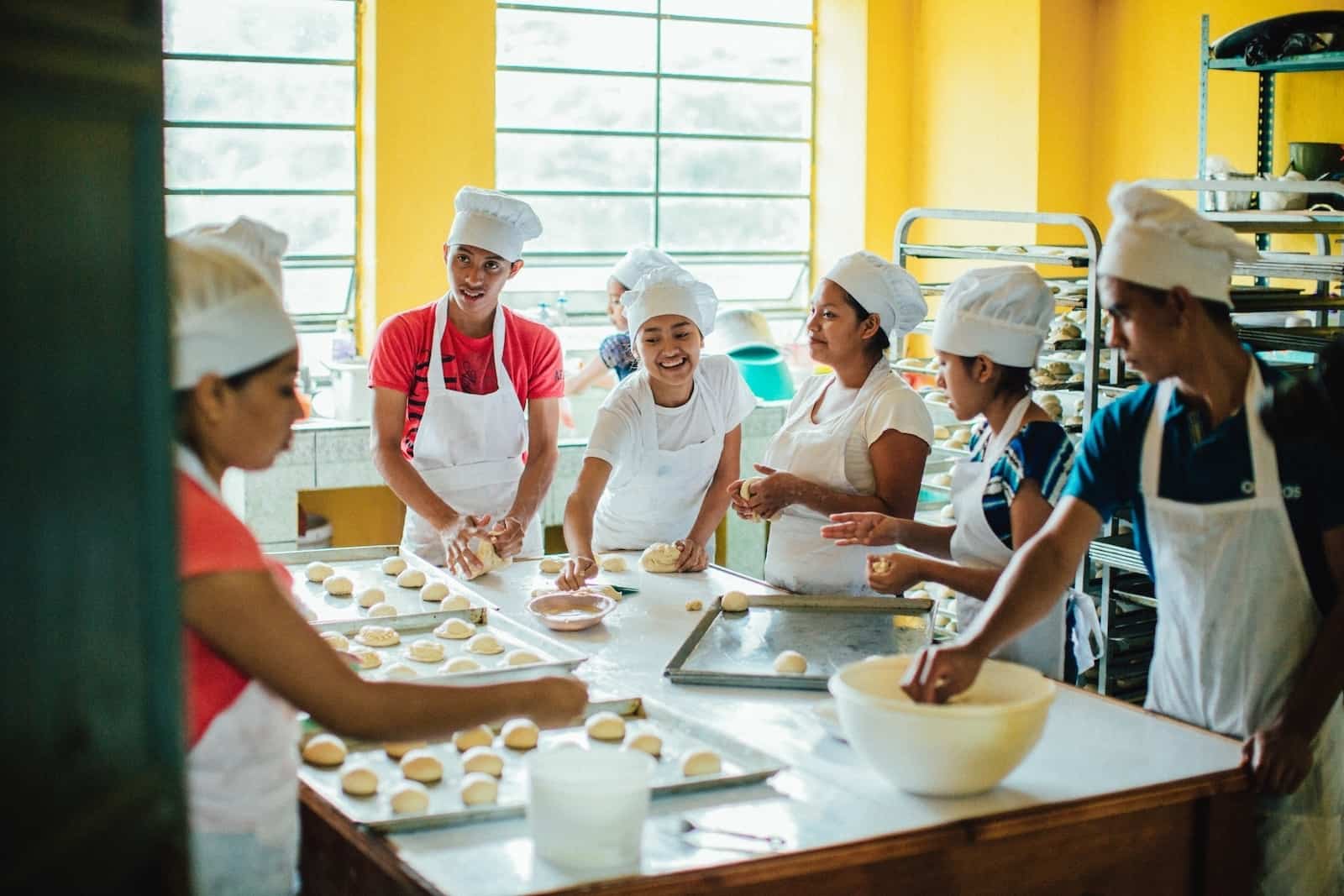 A group of students wear white aprons and chef's hats, standing around a large table, making rolls as they engage in vocational training in baking to defeat poverty. 