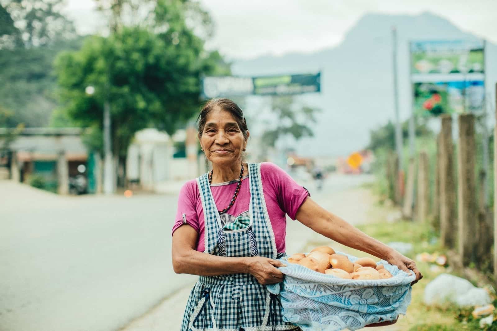 A woman stands outside on a streetside, holding a basket of breads. She wears a blue apron and pink shirt.