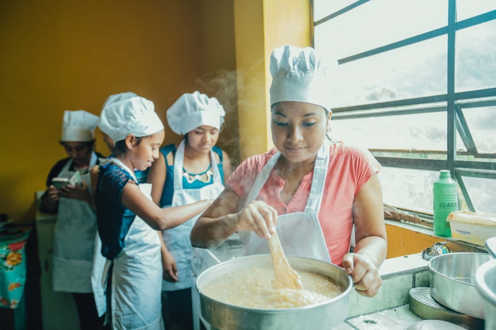 A young woman in a white apron and chef's hat stirs a large pot. Behind her, a group of girls focuses on a pan. They are doing vocational training in baking to escape poverty. 