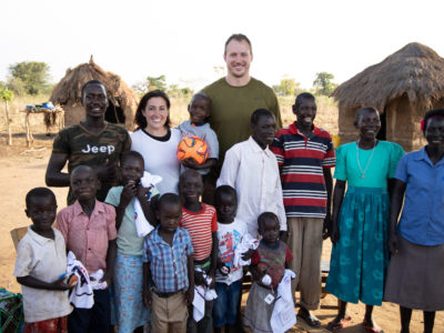 A group of people, Ugandans and two Americans, stand outside with grass thatched huts in the background, everyone smiling at the camera.