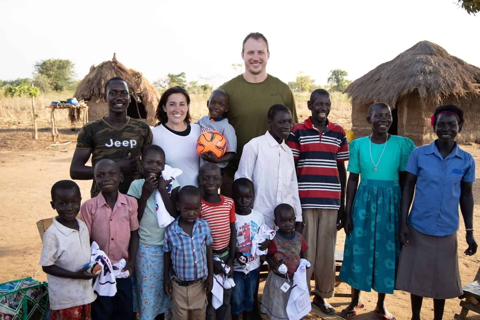 A group of people, Ugandans and two Americans, Nate Solder and his wife, stand outside with grass thatched huts in the background, everyone smiling at the camera.