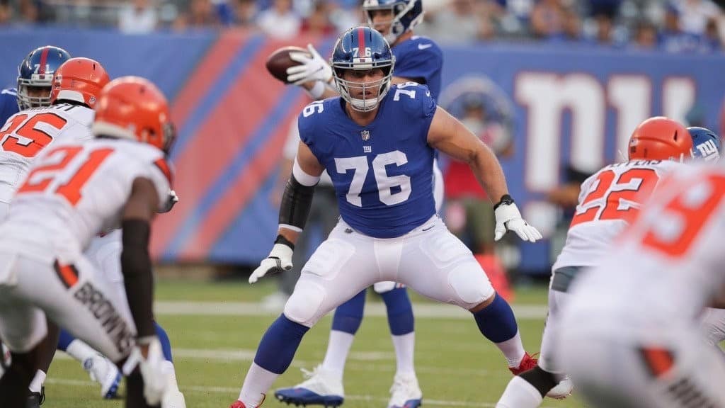 Nate Solder, center, playing football in a blue and white New York Giants uniform. The man behind him holds the ball , while in the foreground men are in white uniforms that say 'Browns.'