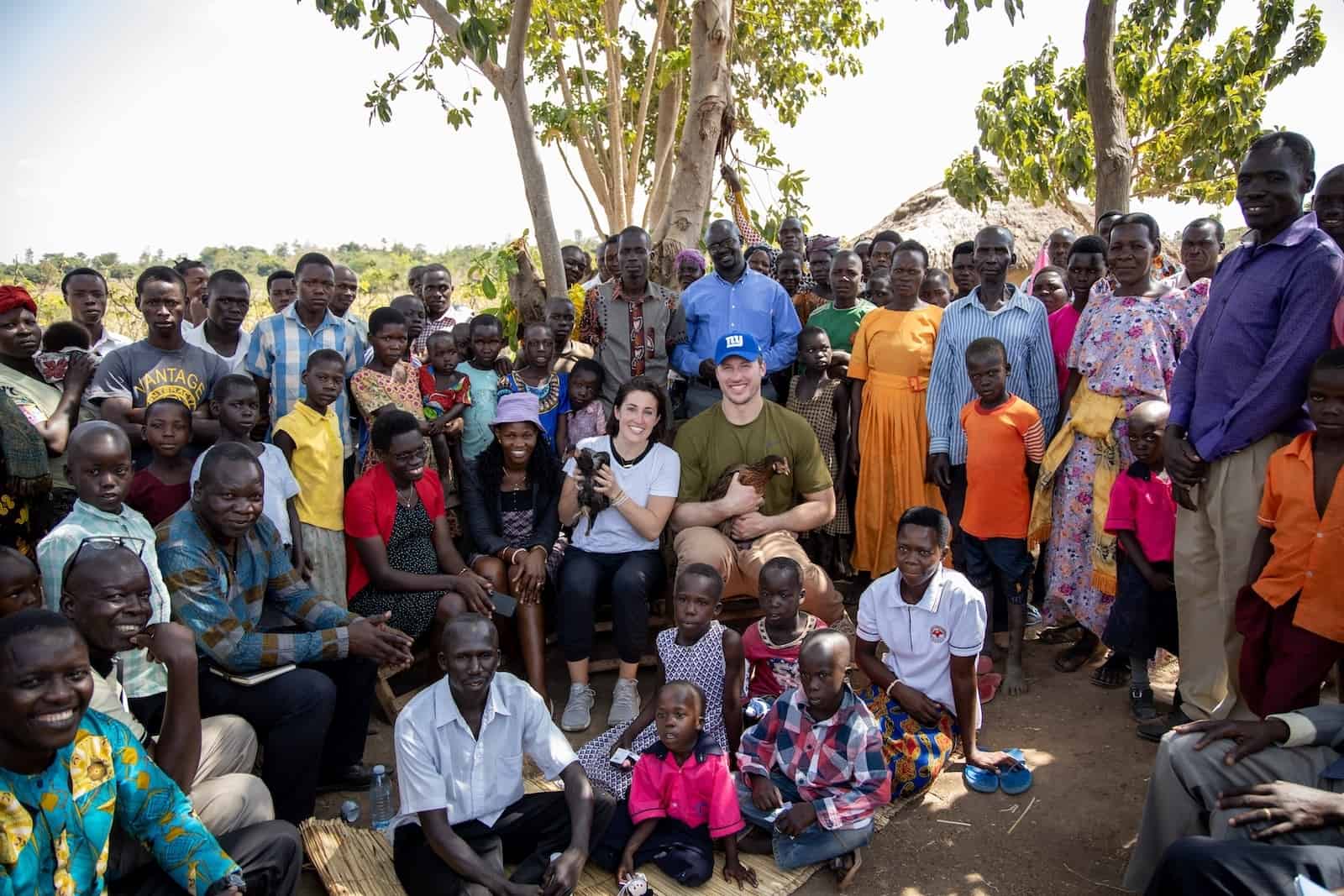 A large group of Ugandans sits and stands outside, posing for a group photo. In the center are two Americans, Nate Solder and his wife, sitting, holding chickens.