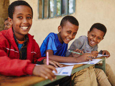 Three boys sit in a row at a desk outside, each writing a letter. They all smile at the camera.